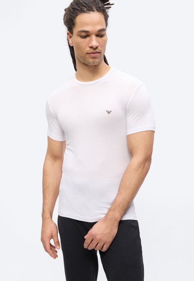 Άνδρας με ντρέντλοκς που φοράει εφαρμοστό λευκό κοντομάνικο πουκάμισο και μαύρο παντελόνι, κοιτάζοντας προς τα κάτω και προς τα αριστερά του.