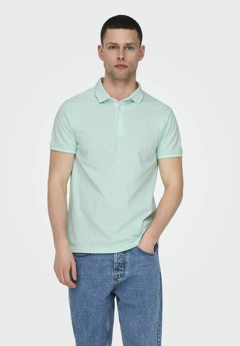 Camisa polo verde claro con cuello, mangas cortas y un pequeño logo. Combinada con jeans azules, demostrando un diseño casual.