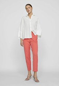 Camicia bianca in lino oversize con chiusura a bottoni, abbinata a pantaloni aderenti color corallo. Modella che indossa sandali con tacco argento.