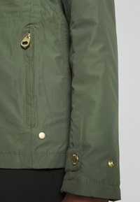 Gros plan de la manche d'une veste verte et de la poche latérale avec curseur de fermeture éclair doré et boutons-pression au poignet et près de la poche.