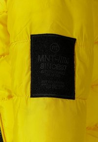 Veste matelassée jaune avec texture en losanges et une étiquette en tissu noire rectangulaire comportant un texte en relief. Design minimaliste.