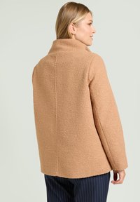 Cappotto in pile fluffy color beige con colletto alto, caratterizzato da una forma boxy e spalle scese. Tessuto testurizzato, dettaglio della cucitura centrale dritta sul retro.