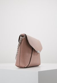 Malá kabelka z prachově růžové kůže trojúhelníkového tvaru, s klopou a stříbrným řetězovým popruhem, umístěná na bílé ploše na světlém pozadí.