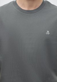 Άνδρας που φοράει απλό γκρι T-shirt με στρογγυλή λαιμόκοψη και μικρά λευκά γράμματα M, O και P τοποθετημένα κάθετα στο αριστερό στήθος.