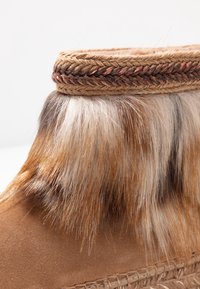 Närbild av en brun mockastövvel med flerfärgad pälskant och flätad detalj nära den övre kanten.