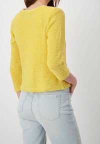 Gul stickad tröja med trekvartslånga ärmar, texturerad yta och rundad halsringning, i kombination med ljusblå denimjeans.