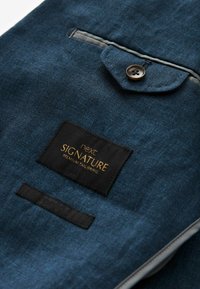 Tamsiai mėlynas audinio švarkas su krūtine kišene su mygtuku, lygiu paviršiumi ir etiketė, kuri rodo "next SIGNATURE PREMIUM TAILORING."