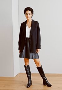 Blazer noir sur un haut blanc, associé à une jupe en cuir noir et des bottes noires jusqu'au genou. Texture lisse, design ajusté.