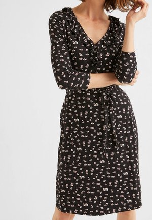 Nainen pukeutuneena mustaan, polvipituiseen mekkoon, jossa on vaaleanpunainen ja valkoinen kukkakuosi, röyhelöinen V-kaulus, 3/4-hihat ja vyötärölle solmittava vyö.