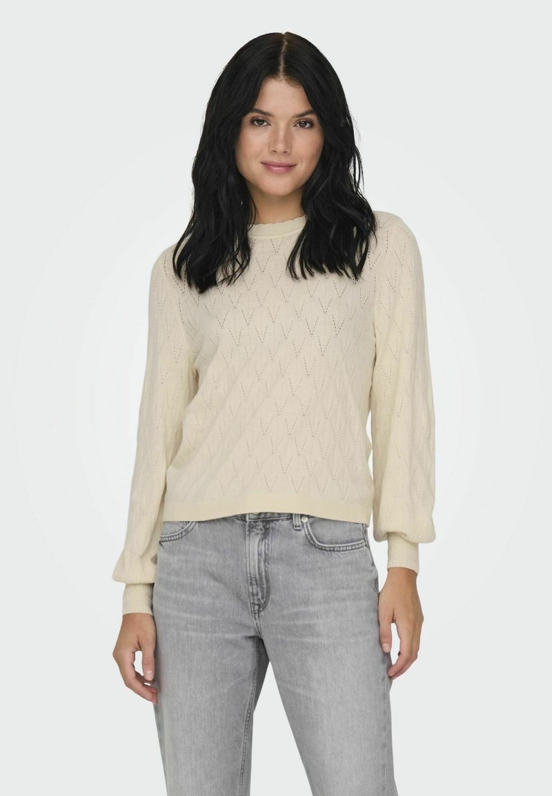 Suéter color crema con patrones en forma de diamante, con un cuello redondo y mangas largas ligeramente abullonadas. Llevado con jeans gris claro.