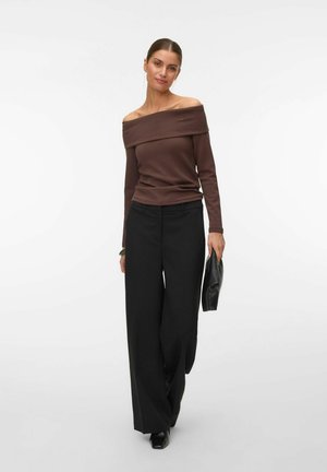 Femme portant un haut marron à épaules dénudées et un pantalon large noir, tenant une pochette noire, debout devant un fond blanc.