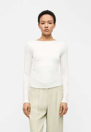 Γυναίκα με κοντά σγουρά μαλλιά, ντυμένη με λευκό εφαρμοστό τοπ μακρυμάνικο και ανοιχτό μπεζ ψηλόμεσο παντελόνι με πιέτες, στέκεται μπροστά σε απλό φόντο.