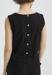 Haut noir sans manches avec cinq boutons au dos, porté par une personne aux cheveux courts et bouclés, assorti à un bas noir et un bracelet rouge.