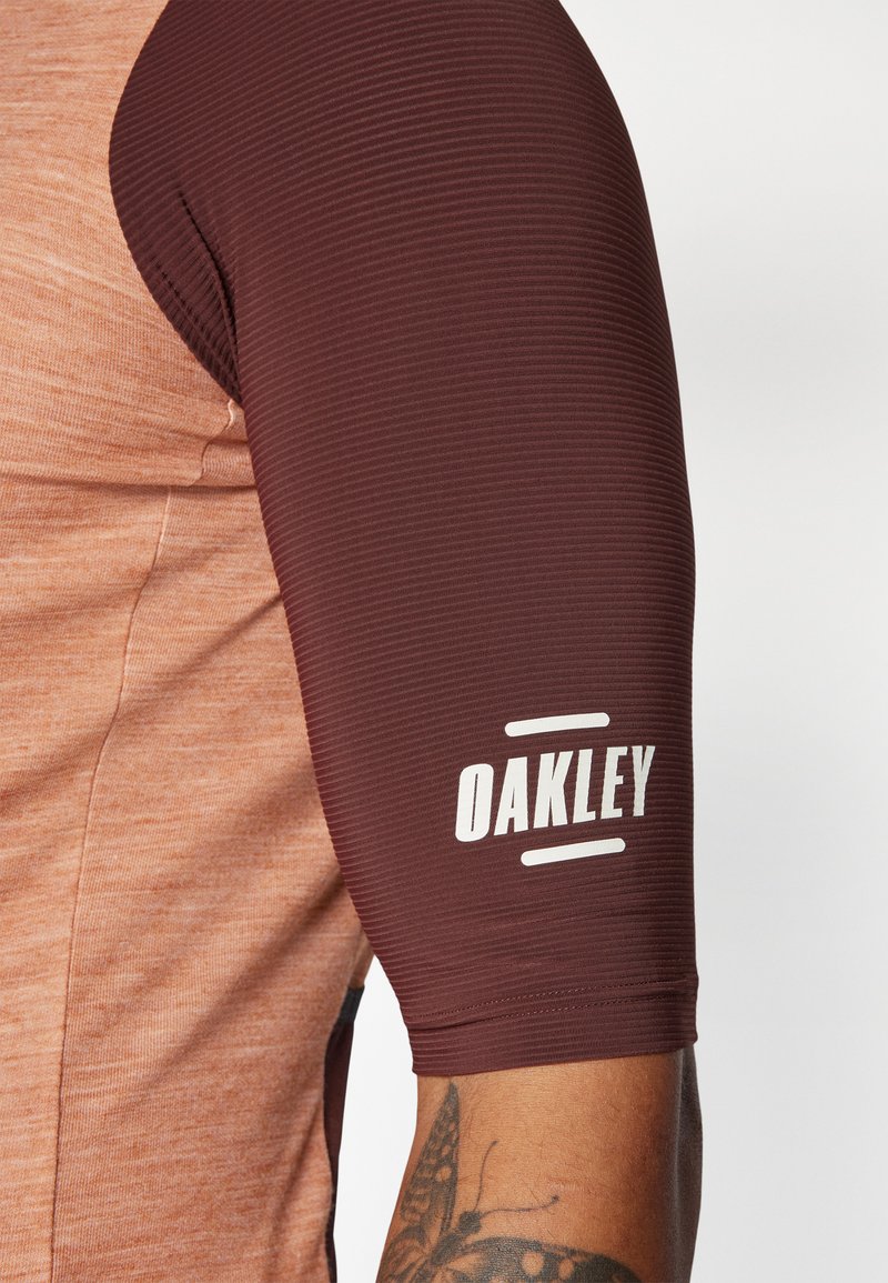 Maglietta atletica a righe rosse e arancioni con maniche corte a coste color bordeaux. Presenta un logo "OAKLEY" bianco sulla parte superiore del braccio. Texture liscia.