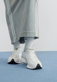 White athletic sneakers with black soles, featuring a textured upper and laces. Worn with light blue ankle socks and a long denim skirt with white stripes.