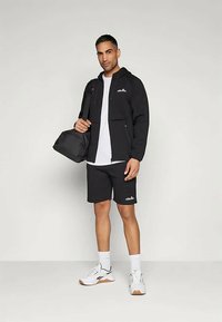 Vyras vilki juodą užtrauktuku užsegamą džemperį, juodas šortus, baltą marškinėlį ir baltus sportbačius, ant peties laiko juodą sportinę kuprinę, stovi.