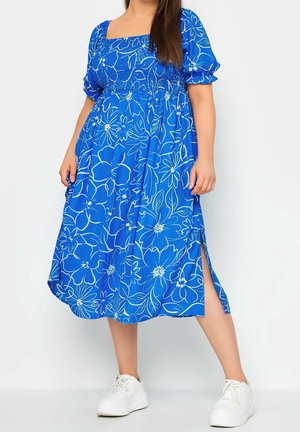 Vrouw draagt een blauwe midi-jurk met bloemenprint, pofmouwen en een zijsplit, gecombineerd met witte sneakers, staand tegen een witte achtergrond.