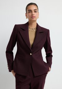 Femme portant un costume ajusté bordeaux foncé et un top beige avec des embellissements, standing avec les mains dans les poches sur un fond uni.