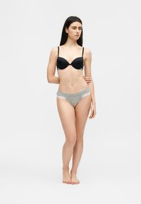 Γυναίκα όρθια, ξυπόλυτη, με μαύρο σουτιέν και γκρι εσώρουχο με δαντέλα, μπροστά σε απλό φωτεινό φόντο.