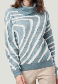 Frau in einem blauen Rollkragenpullover mit weißen abstrakten Streifen und beigen Hosen, die vor einem schlichten hellen Hintergrund steht.