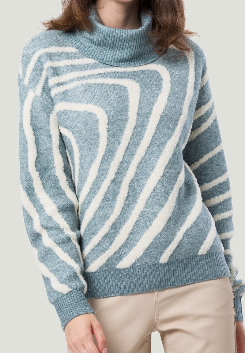 Frau in einem blauen Rollkragenpullover mit weißen abstrakten Streifen und beigen Hosen, die vor einem schlichten hellen Hintergrund steht.