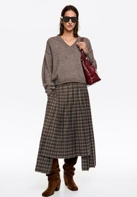 Brązowy dziergany sweter z dekoltem w serek, zestawiony z plisowaną spódnicą w brązowej tonacji. Modelka trzyma małą bordową skórzaną torebkę. Brązowe kozaki za kolano.