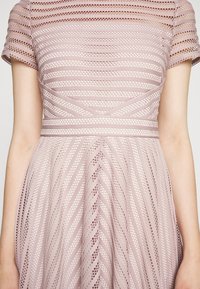 Jasnoróżowa sukienka z poziomymi i diagonalnymi otwartymi wzorami dzianiny, krótkimi rękawami oraz dopasowanym stanem i rozszerzanym dołem.