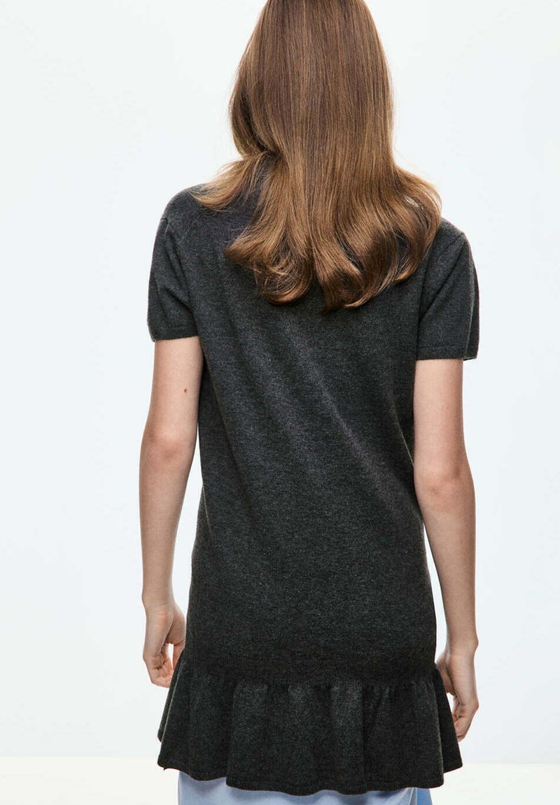 Femme aux cheveux châtain clair portant une robe gris foncé à manches courtes avec un ourlet à volants, debout, tournée de dos sur un fond uni.