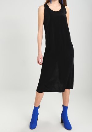 Vestito midi nero con scollatura a barca e design senza maniche. Tessuto morbido, indossato con stivaletti blu alla caviglia. Silhouette semplice e dettagli minimi.