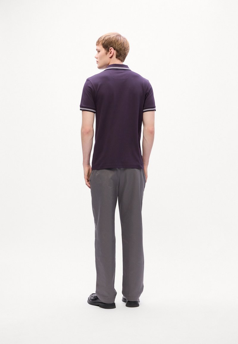 Un jeune homme de dos portant un polo violet foncé avec des bordures blanches et un pantalon gris, sur fond blanc.