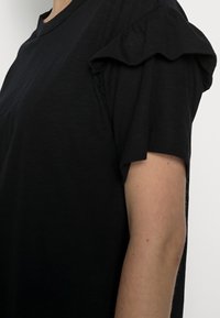 Schwarzes T-Shirt mit kurzen Ärmeln und Rüschendetail an der Schulter. Der Stoff wirkt weich mit einer subtilen Textur. Standard Rundhalsausschnitt.