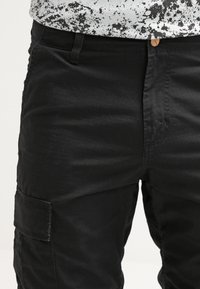 Pantalons cargo noirs avec une texture lisse, poche latérale et un design ajusté. Fermeture à la taille avec un petit bouton circulaire.