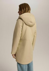Beżowa kurtka z kapturem o prostym kroju, z gładką strukturą i dłuższym krojem. Jasnoniebieskie dżinsy dopełniają strój.