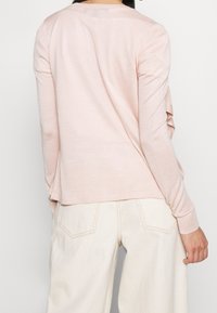 Světle růžový top s dlouhými rukávy vyrobený z měkké tkaniny, s uvolněným střihem a volánkovými boky a zaobleným výstřihem. V kombinaci s béžovými širokými kalhotami.