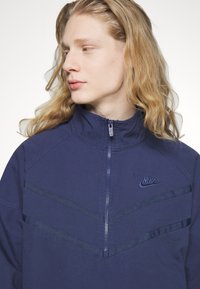 Jūras spēku krāsas zip-up jaka ar augstu apkakli, ar diogonālām svītrām un nelielu Nike logo uz krūts. Izgatavota no teksturēta auduma.