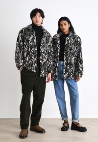 Zwart-wit gestreepte fleecjassen op twee modellen. De ene draagt deze in combinatie met donkergroene corduroy-broeken, de andere met lichtblauwe jeans en zwarte schoenen.