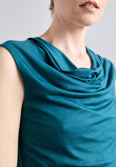 Nærbilde av en person som har på seg en ermeløs tåkeblå topp med drapert hals, som viser nakken, leppene og øvre bryst.