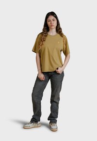 Camiseta de algodón color verde oliva con cuello redondo y mangas cortas, que presenta un pequeño logotipo naranja. Combinada con jeans oscuros y zapatillas deportivas.