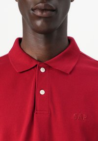 Punane polo särk tekstuuriga kangast, klassikalise kaelusega, kolme nupuga资料ja ja tikitud "GAP" logo samas värvis.