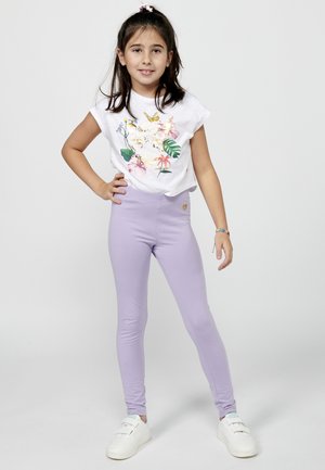 Camiseta blanca de manga corta con diseño floral; leggings lavanda; zapatillas blancas; chica posa con una mano en la cadera, sonriendo.