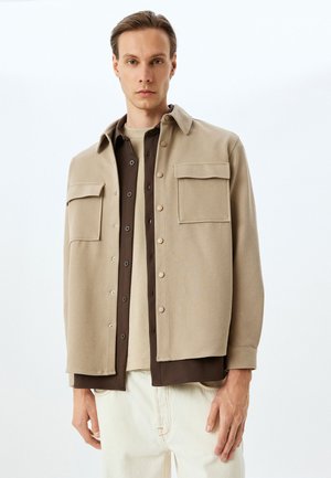 Beige jakke med krave og to brystlommer, lukket med trykknapper, båret over en brun skjorte og en kremfarvet top, parret med lyse bukser.