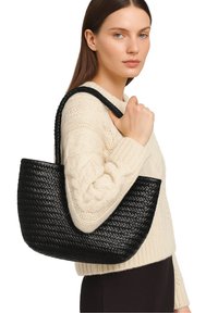 Donna che indossa un maglione a costine color crema, mentre tiene una borsa a tracolla in pelle intrecciata nera su uno sfondo bianco.