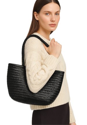 Donna che indossa un maglione a costine color crema, mentre tiene una borsa a tracolla in pelle intrecciata nera su uno sfondo bianco.