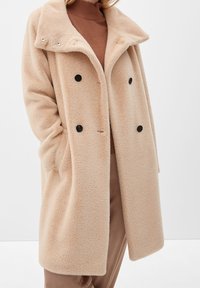 Kvinna som bär en beige, dubbelknäppt luddig kappa över en brun tröja och ljusbruna byxor, stående med händerna i kappfickorna.