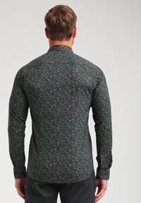 Majica s dolgimi rokavi v temno zeleni tkanini z ponavljajočim se svetlim listnatim vzorcem, ki ima klasičen ovratnik in zapnjen manšeti.