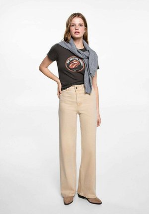 Bež traperice širokog kroja s visokim strukom, uparene s tamnom majicom s grafikom. Sivi pulover prebačen preko ramena. Glatka tekstura tkanine.