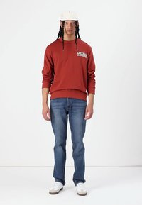 Roestkleurige sweatshirt met 'TIMBS' logo op de borst, gecombineerd met blauwe jeans en witte sneakers. Model heeft vlechten en een pet.