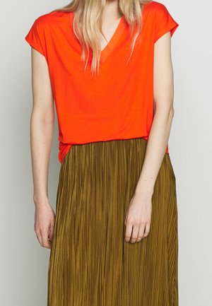 Femme portant un haut orange vif à manches courtes et col en V, ainsi qu'une jupe plissée taille haute vert olive, se tenant devant un fond uni.