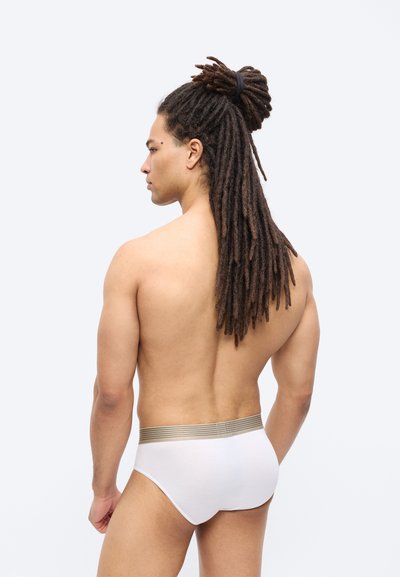 Άνδρας με μακριά dreadlocks δεμένα πίσω, γυμνό πάνω μέρος σώματος, φοράει λευκά εσώρουχα με μπεζ ελαστική μέση, στραμμένος προς τα αριστερά σε απλό φόντο.