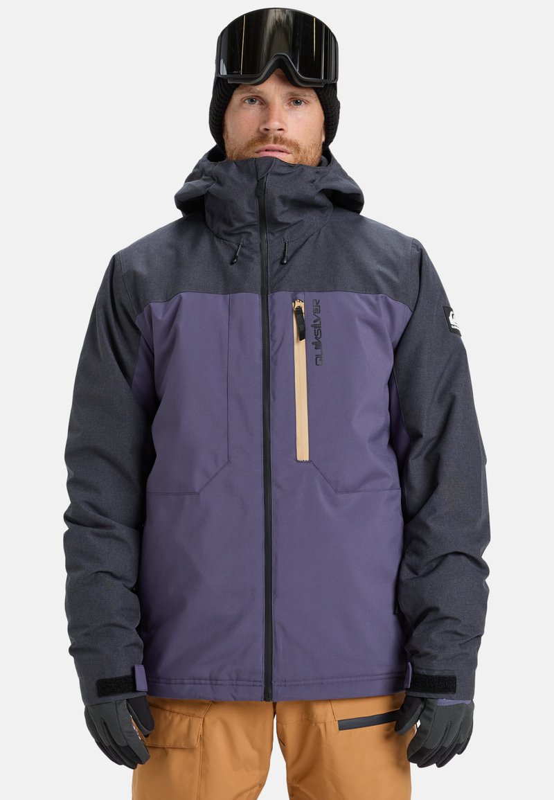 Man wearing a two-tone purple and dark gray winter jacket, mustard pants, black gloves, black beanie, and ski goggles on forehead.